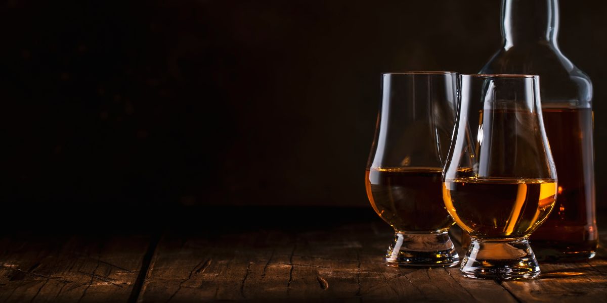 Two whiskey glasses filled with bourbon next to a bottle on a wooden surface against a dark background.