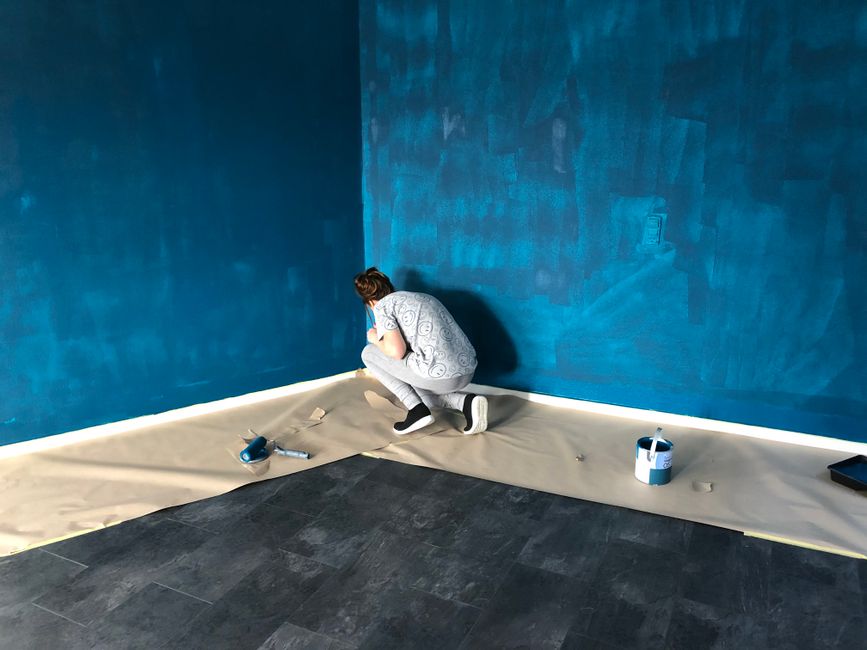 A focused individual on a stepladder carefully painting a living room wall with a roller in a bright, neutral beige color to prepare a home for sale.